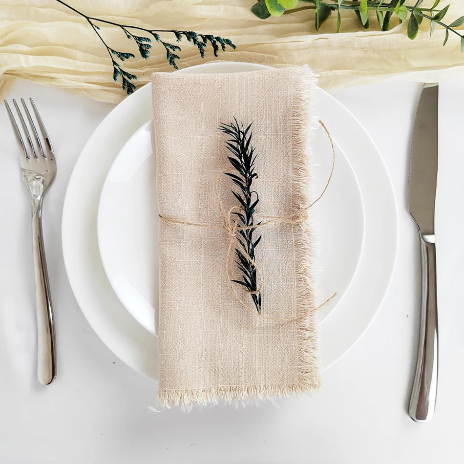 Une serviette de table en lin beige avec un motif de feuilles découpées de manière élégante, entourée de couverts sur une assiette blanche et de branches de plantes vertes.