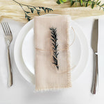 Une serviette de table en lin beige avec un motif de feuilles découpées de manière élégante, entourée de couverts sur une assiette blanche et de branches de plantes vertes.