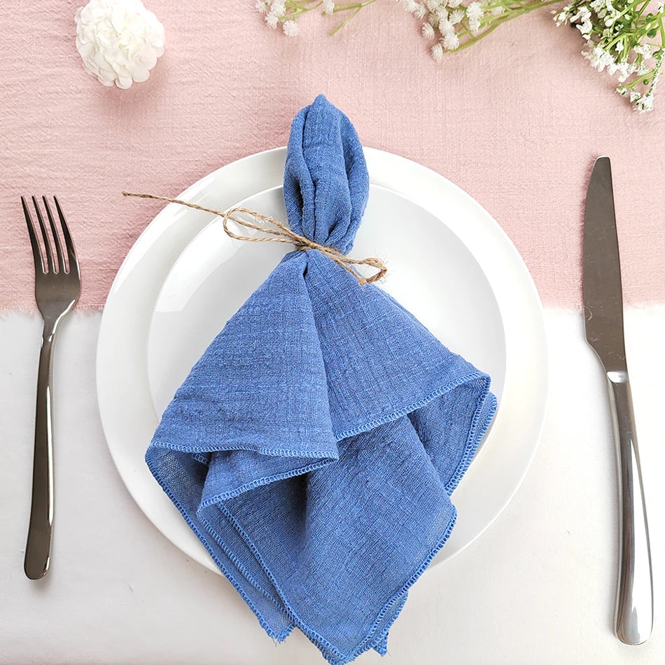 Une serviette de table en lin bleu marine enroulée sur une assiette blanche avec des couverts sur un fond rose pastel.