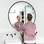 Un homme souriant regarde son reflet dans un miroir rond noir dans une salle de bain lumineuse et épurée.