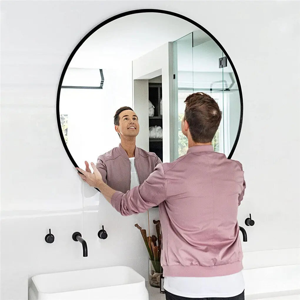 Un homme souriant regarde son reflet dans un miroir rond noir dans une salle de bain lumineuse et épurée.