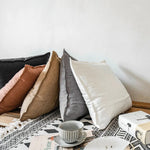 Pile de coussins décoratifs aux couleurs neutres sur un tapis aux motifs ethniques, avec une tasse et une soucoupe en céramique blanche posées sur le sol.