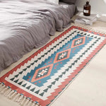 Tapis géométrique multicolore de style aztèque avec des motifs triangulaires et un jeu de couleurs chaudes et froides, posé au pied du lit.
