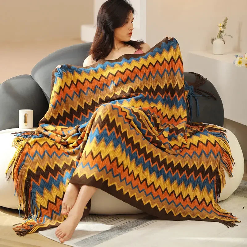 Une femme enveloppée dans une couverture tricotée en zigzag multicolore aux nuances chaudes, assise confortablement sur un fauteuil.