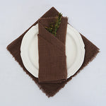 Une serviette de table en tissu brun foncé, pliée avec soin sur une assiette blanche, est décorée de quelques brins de verdure.