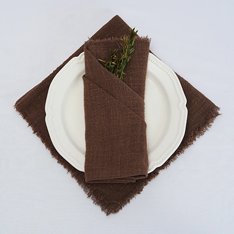 Une serviette de table en tissu brun foncé, pliée avec soin sur une assiette blanche, est décorée de quelques brins de verdure.