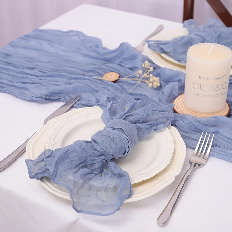 Sur une table blanche, on peut voir une assiette blanche avec une serviette en tissu bleu clair posée dessus. Près de l'assiette se trouve une bougie blanche dans un support en bois, ainsi que quelques petites fleurs blanches. L'ensemble donne une ambiance calme et sereine.