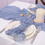 Sur une table blanche, on peut voir une assiette blanche avec une serviette en tissu bleu clair posée dessus. Près de l'assiette se trouve une bougie blanche dans un support en bois, ainsi que quelques petites fleurs blanches. L'ensemble donne une ambiance calme et sereine.