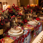 Décor de table de Noël festif avec des branches de sapin, des bougies allumées, des verres, des plats et des décorations rouges et dorées, disposés sur une nappe à motifs colorés.