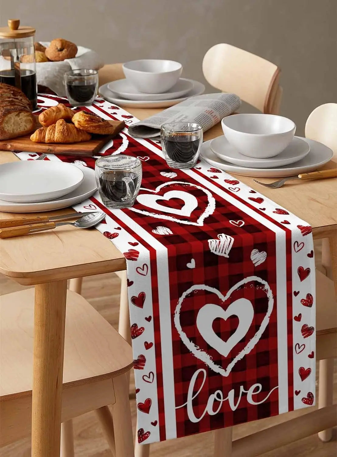 Une table dressée avec des assiettes, des verres et des pâtisseries, le tout surplombé d'un chemin de table rouge décoré de coeurs blancs et du mot "love".