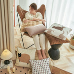 Jeune femme assise confortablement dans un fauteuil cosy, enveloppée dans un plaid moelleux, entourée de coussins décoratifs et d'accessoires de style scandinave, créant une atmosphère chaleureuse et accueillante.