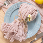 Une serviette froissée en tissu de couleur rose-gris, nouée avec de la ficelle, et décorée d'une petite fleur blanche, repose sur une assiette gris bleu posée sur une surface en bois clair.