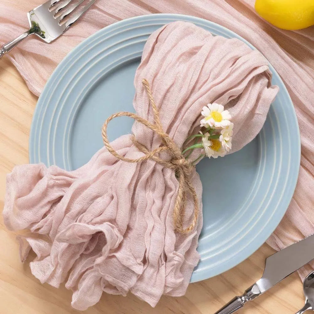Une serviette froissée en tissu de couleur rose-gris, nouée avec de la ficelle, et décorée d'une petite fleur blanche, repose sur une assiette gris bleu posée sur une surface en bois clair.