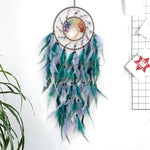 Un attrape-rêves décoratif composé d'un anneau entouré de plumes vertes, bleues et grises, avec un soleil multicolore en son centre, suspendu par un fil.
