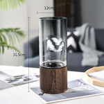 Vase décoratif en verre transparent sur socle en bois sombre, mesurant 31,5 cm de hauteur et 12 cm de diamètre, posé sur une surface texturée avec des accents végétaux en arrière-plan.