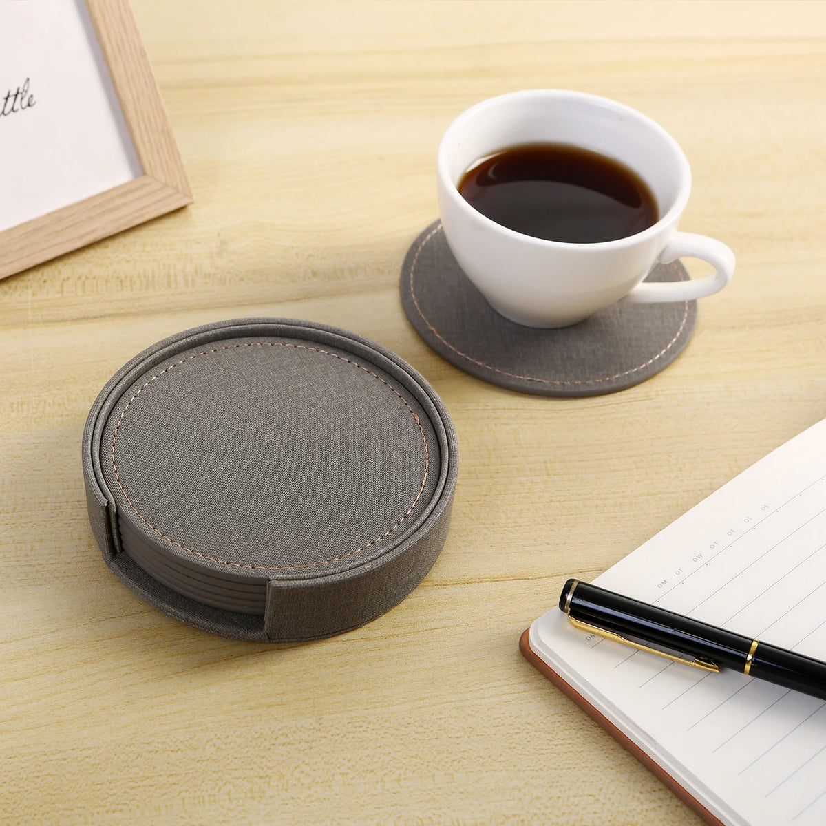 Une tasse de café, des sous-verres gris en tissu et un stylo sur un bureau en bois avec un cadre photo, prêts pour une pause de travail relaxante.