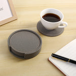 Une tasse de café, des sous-verres gris en tissu et un stylo sur un bureau en bois avec un cadre photo, prêts pour une pause de travail relaxante.