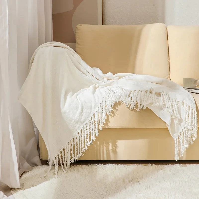 Couverture blanche douce et texturée avec des franges ornementales sur un canapé beige dans un environnement intérieur chaleureux et confortable.