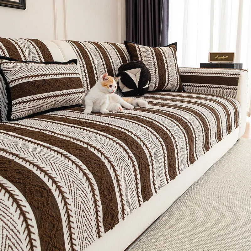 Un chat blanc mignon et petit est assis confortablement sur un canapé marron et beige avec un motif géométrique, dans une pièce bien éclairée.