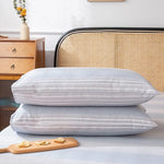 Ensemble de draps de lit rayés dans des tons gris et bleus, posés sur un lit avec une tête de lit en osier, dans une chambre confortable et douillette.