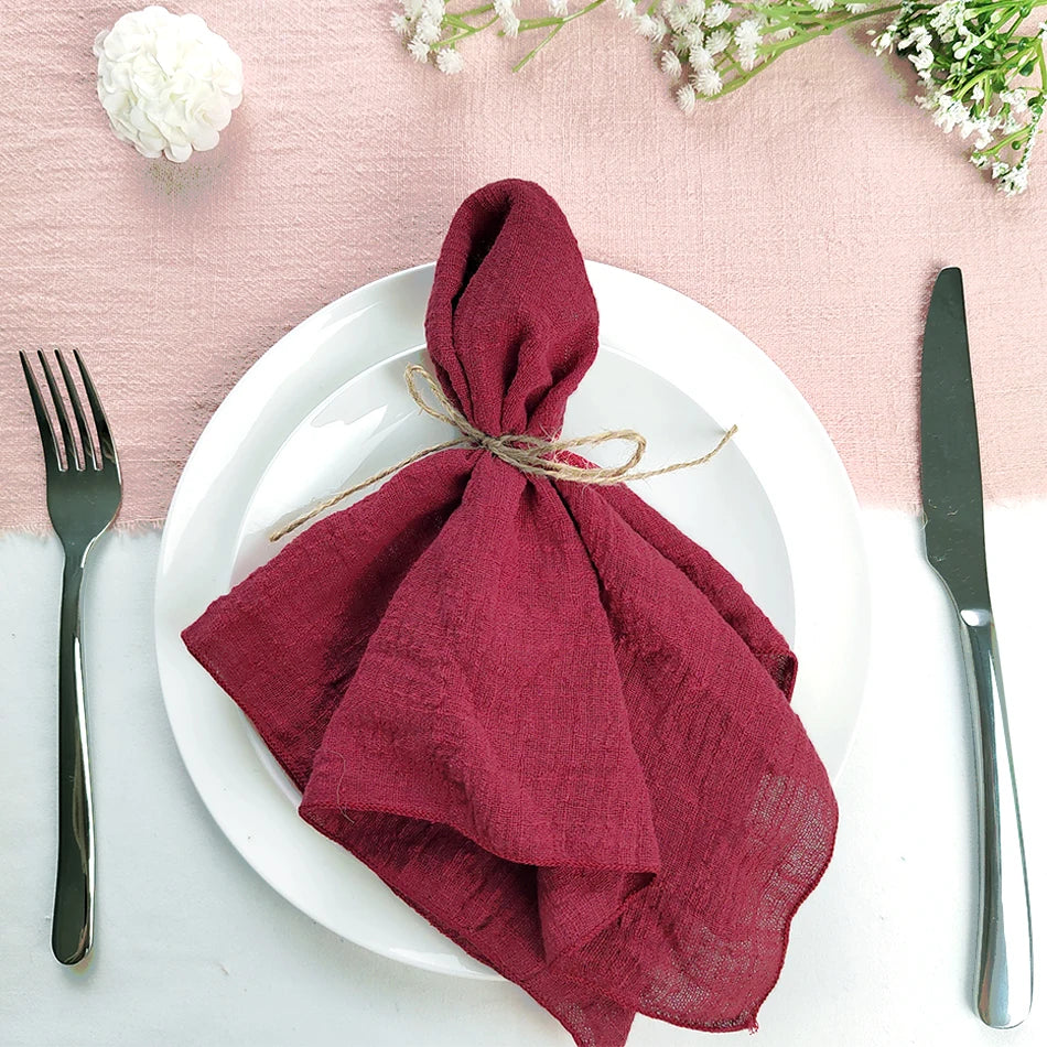 Une nappe blanche avec des fleurs blanches et une serviette en tissu bordeaux roulée et attachée avec une ficelle sur une assiette blanche.