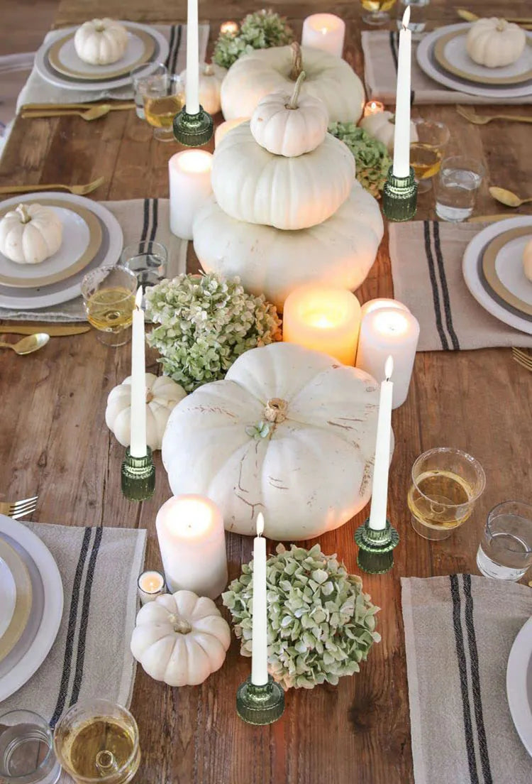 Cette image présente une magnifique table d'automne décorée de citrouilles blanches, de fleurs d'hortensia, de chandelles et de verres de vin, créant une ambiance chaleureuse et festive.
