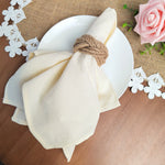 Sur un fond de jute brun, on voit une assiette blanche avec un linge blanc plié et noué avec de la corde de jute, entouré de fleurs blanches en papier et d'une rose rose.