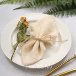 Une belle décoration de table avec un sachet décoratif en lin et feuillage sur un plat blanc avec des couverts dorés.