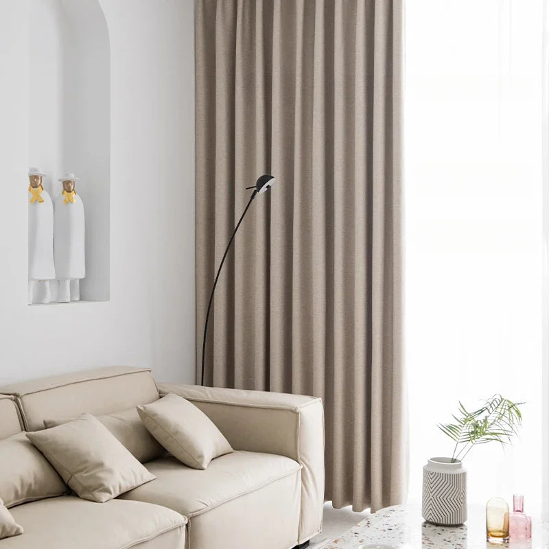 Salle de séjour moderne avec rideaux beiges offrant une ambiance chaleureuse. Canapé confortable assorti avec coussins pour un confort optimal. Décoration minimaliste avec des statuettes et des plantes d'intérieur, apportant une touche de fraîcheur et d'élégance à la pièce. Parfaite harmonie pour un intérieur contemporain et accueillant.