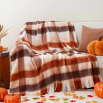 Couverture douillette à carreaux oranges et bruns sur un canapé, entourée de citrouilles et de feuilles d'automne, créant une ambiance chaleureuse et conviviale.