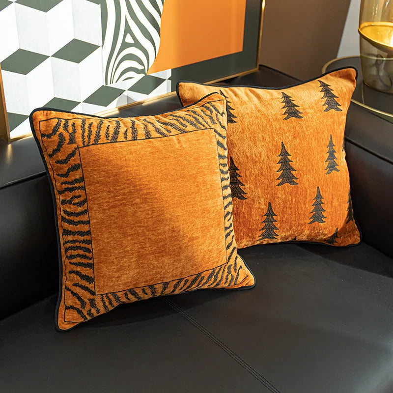 Deux oreillers décoratifs sur un canapé, l'un avec un motif de fourrure de tigre et l'autre avec un motif de sapins noirs sur fond orangé, créant une ambiance chaleureuse et élégante dans l'intérieur.