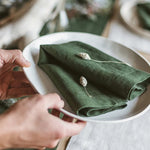 Deux mains disposent délicatement une étoffe verte sur une assiette blanche, avec quelques petites fleurs blanches posées délicatement sur le tissu.