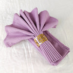 Cette image montre un éventail de serviettes de table en tissu violet, pliées avec élégance et maintenues ensemble par un anneau décoratif en métal doré.