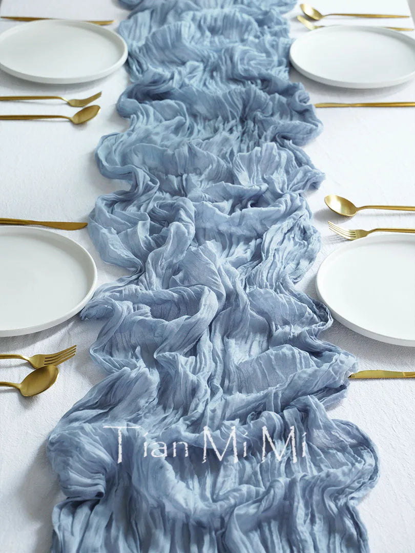 L'image montre un chemin de table en tissu froissé et drapé dans des tons de gris avec des couverts en métal doré disposés sur une table blanche, créant une atmosphère élégante et sophistiquée.