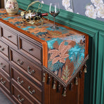 La photo montre une commode en bois avec un chemin de table motif tropical coloré, décoré d'une bouteille de vin et d'un verre à vin posés sur le dessus.