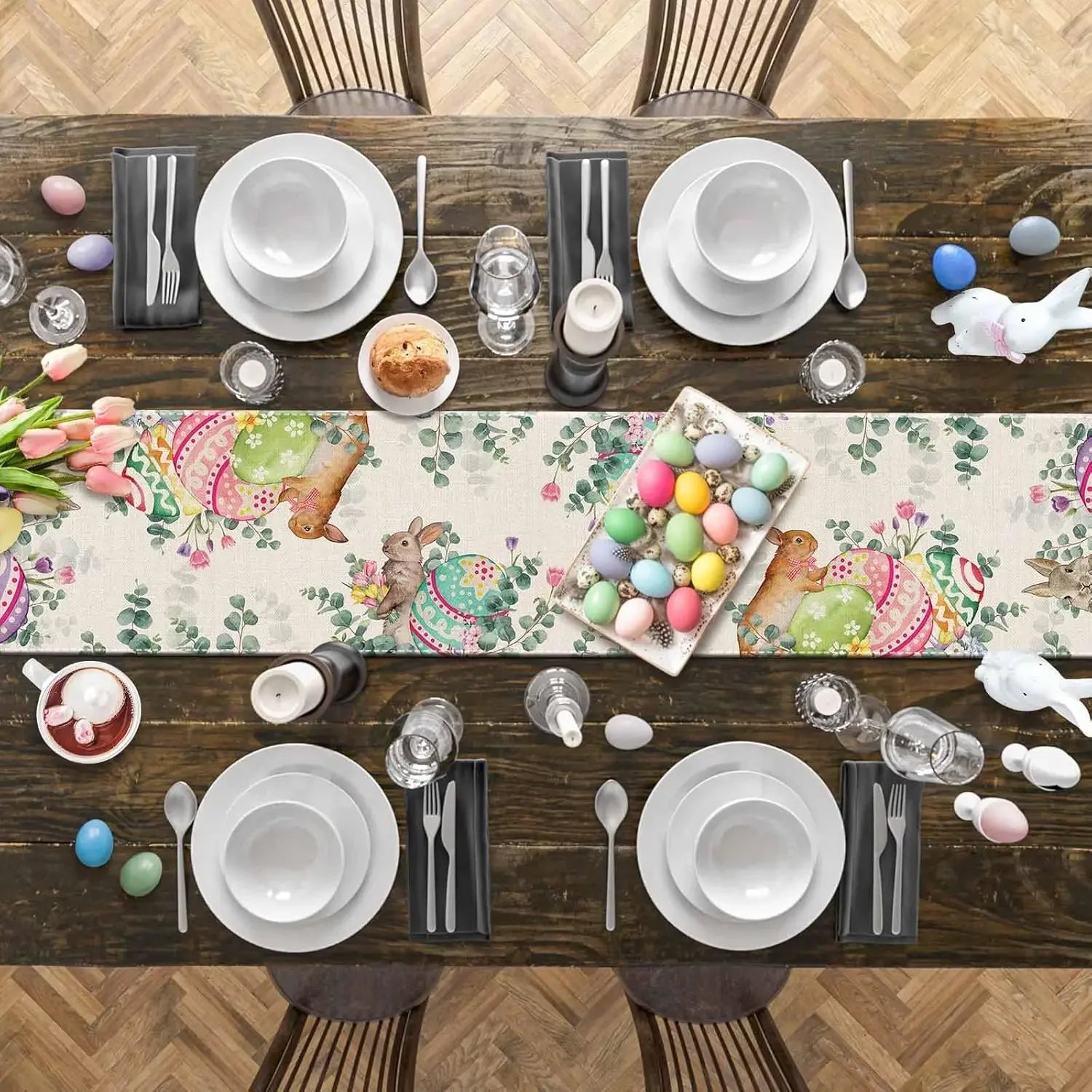 Une table joliment décorée avec des assiettes, des couverts, des ornements de Pâques et un chemin de table coloré représentant des lapins et des œufs.