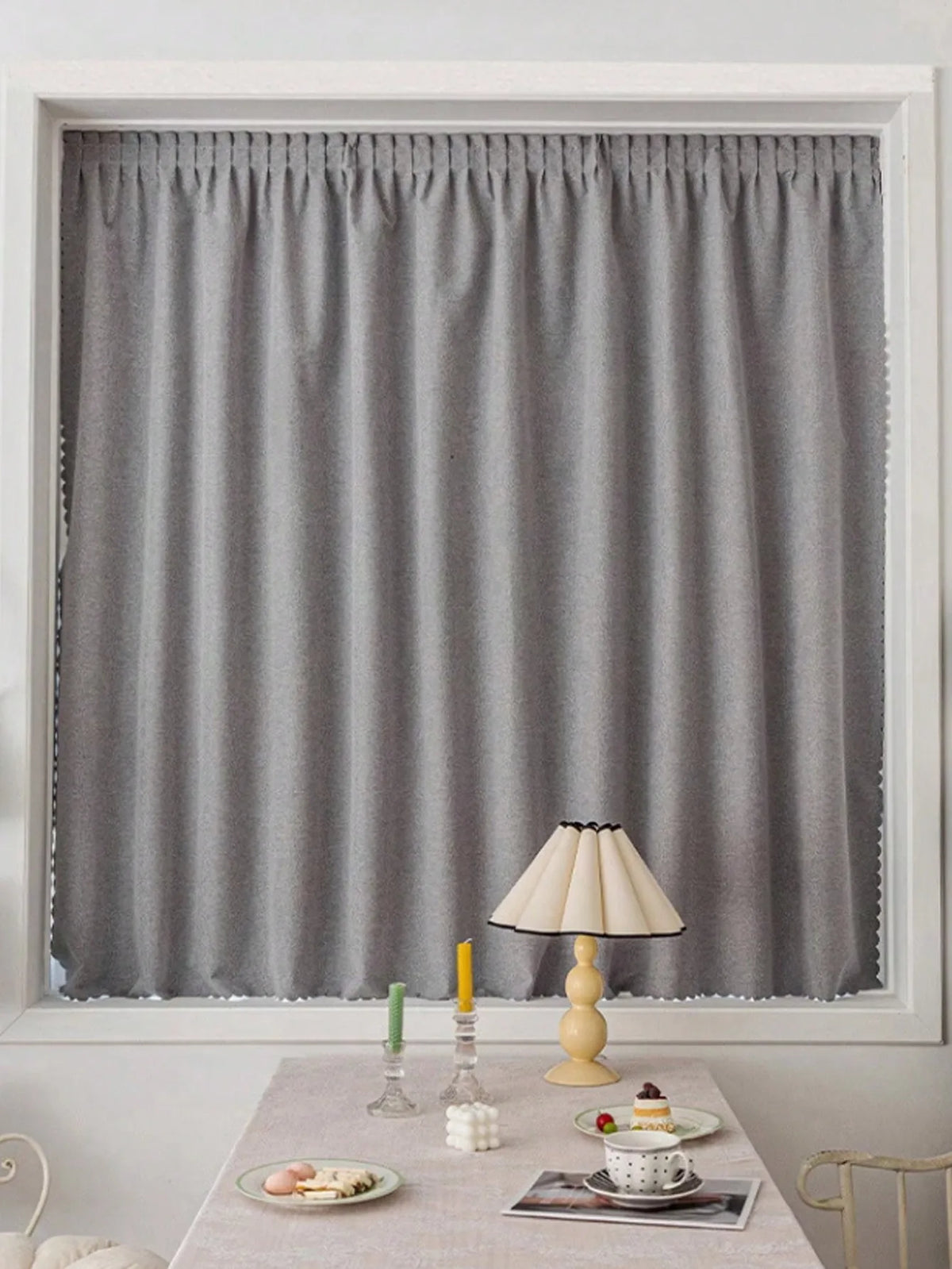 Rideaux modernes gris pour salon, élégamment accrochés dans une fenêtre, offrant une ambiance chaleureuse et tamisée. Idéal pour ajouter une touche de sophistication à votre intérieur. Accompagné d'une table décorée avec une lampe et des accessoires de table colorés. Solution parfaite pour créer un espace de vie accueillant et stylé.