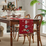 Une table en bois richement décorée avec une nappe rouge ornée de motifs hivernaux, entourée de chaises et de décorations de Noël, dans un intérieur cosy et chaleureux.