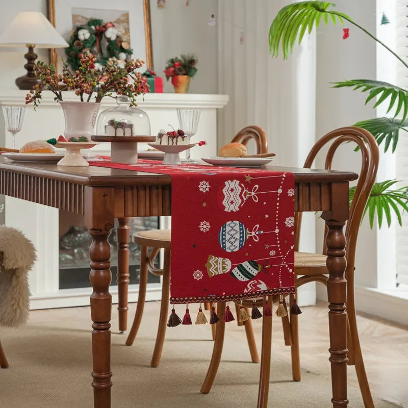 Une table en bois richement décorée avec une nappe rouge ornée de motifs hivernaux, entourée de chaises et de décorations de Noël, dans un intérieur cosy et chaleureux.