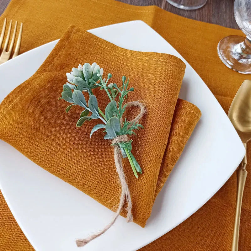 Une serviette de table en lin de couleur ocre, ornée d'un bouquet de fleurs vertes attaché avec une ficelle, sur une assiette blanche.