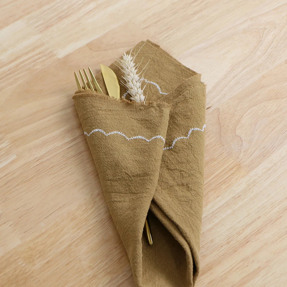 Un élégant rouleau de serviette de table en lin kaki, avec un motif ondulé blanc, contenant des couverts dorés.