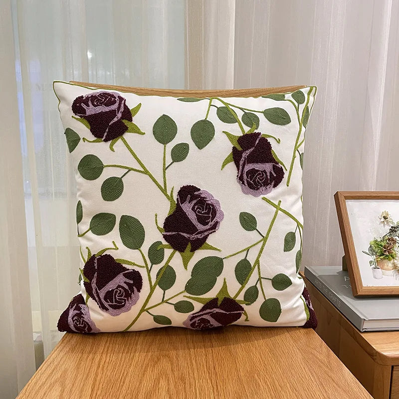 Coussin décoratif avec un motif élégant de roses pourpres et de feuilles vertes sur un fond beige, créant une composition florale apaisante.