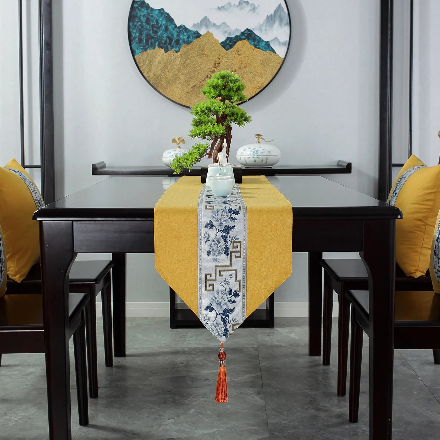 Cette image montre une table à manger dans un intérieur moderne et épuré, avec un chemin de table jaune vif et un superbe décor asiatique comprenant un bonsaï, des vases et une œuvre d'art murale représentant des montagnes brumeuses.