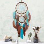 Attrape-rêves rustique décoré de plumes colorées, avec des perles et un design en forme d'araignée, suspendu sur un mur blanc avec des éléments décoratifs.