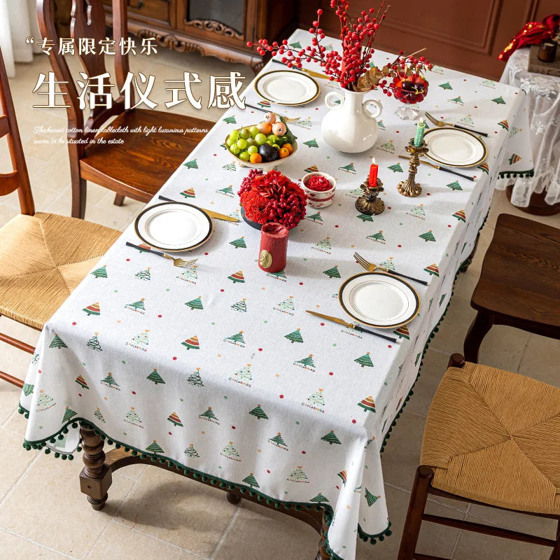 Sur cette image, une décoration de table de fête est visible. La table est recouverte d'une nappe blanche avec des motifs d'arbres de Noël verts et rouges. Des assiettes, des couverts et un arrangement floral composé de fleurs rouges et de baies ornent la table.