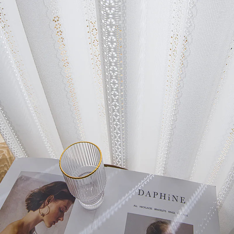 Gros plan sur le voilage blanc brodé posé devant une table décorée d’un verre et d’un magazine.