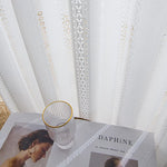 Gros plan sur le voilage blanc brodé posé devant une table décorée d’un verre et d’un magazine.