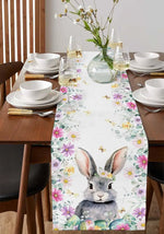 Une table de salle à manger joliment dressée avec des fleurs et des verres de vin, dotée d'une nappe décorée de motifs de fleurs colorées et d'un adorable lapin au centre.