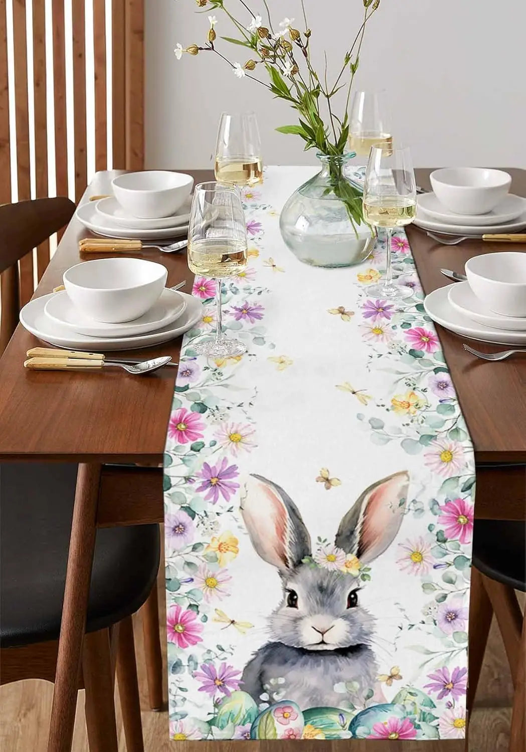 Une table de salle à manger joliment dressée avec des fleurs et des verres de vin, dotée d'une nappe décorée de motifs de fleurs colorées et d'un adorable lapin au centre.