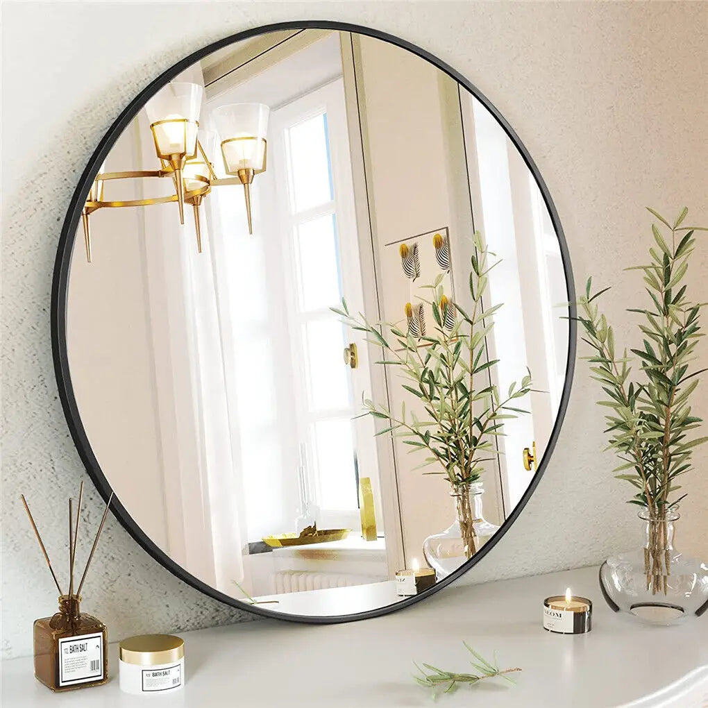 Un grand miroir rond avec un cadre noir décoré d'appliques dorées et entouré de plantes vertes, reflétant une fenêtre lumineuse et une décoration intérieure élégante.